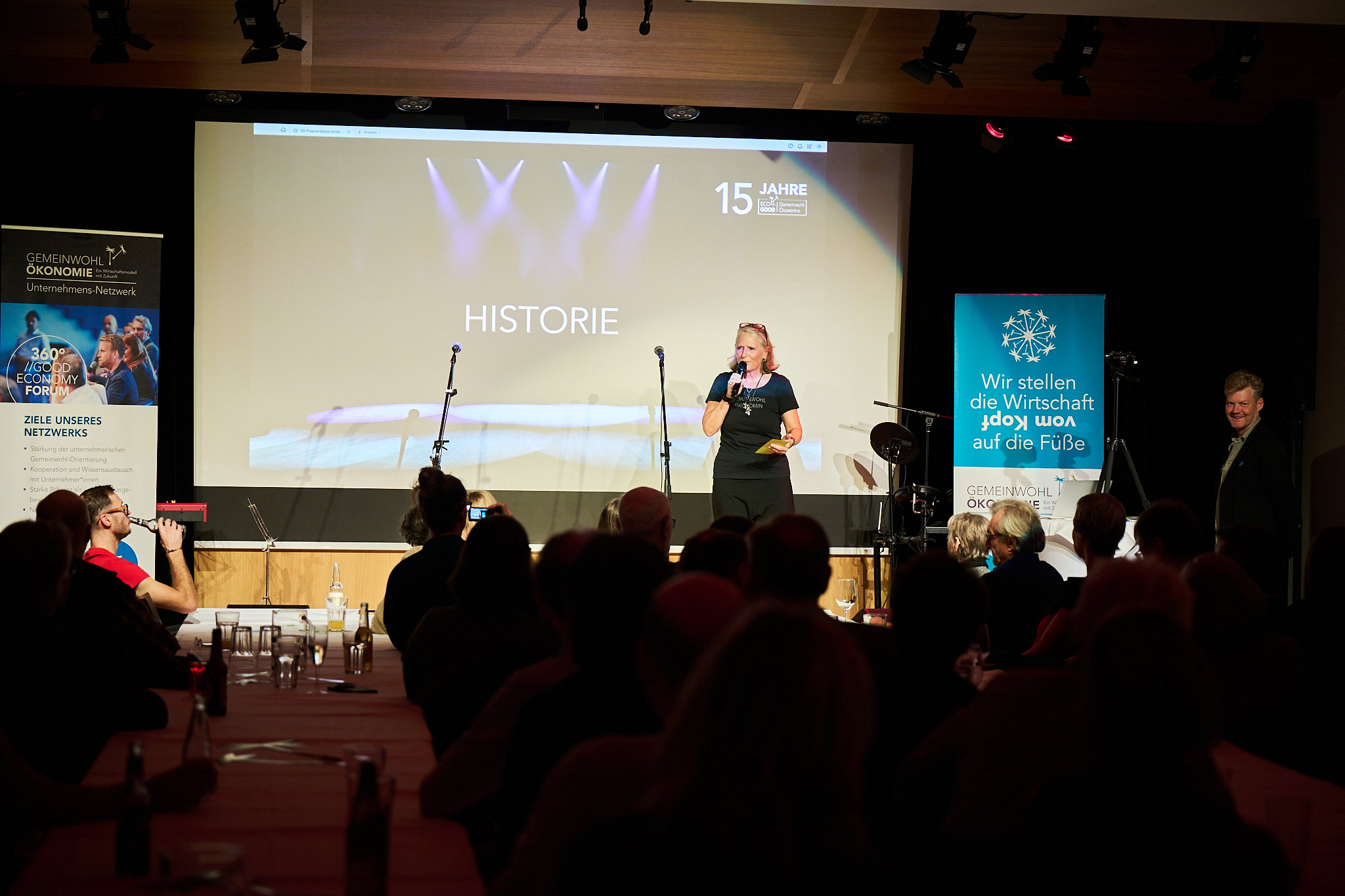 A speaker on stage addresses an audience at an event. The backdrop displays 15 Jahre and Historie. Two banners with text are on either side of the stage.